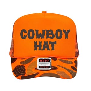 Puede incluir: Gorra de camionero naranja con las palabras "COWBOY HAT" en letras oscuras grises. El panel frontal es naranja y la visera presenta un patr&oacute;n de camuflaje en tonos marrones y naranjas. La gorra tiene una parte trasera de malla y un cierre a presi&oacute;n.