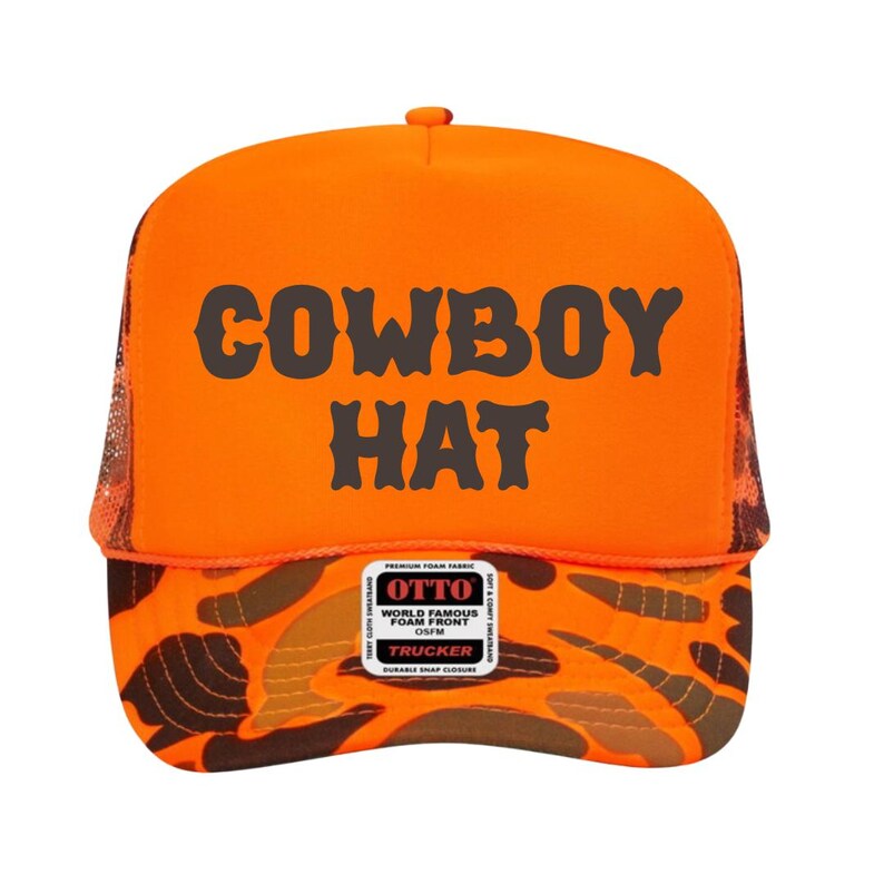 Puede incluir: Gorra de camionero naranja con las palabras "COWBOY HAT" en letras oscuras grises. El panel frontal es naranja y la visera presenta un patr&oacute;n de camuflaje en tonos marrones y naranjas. La gorra tiene una parte trasera de malla y un cierre a presi&oacute;n.