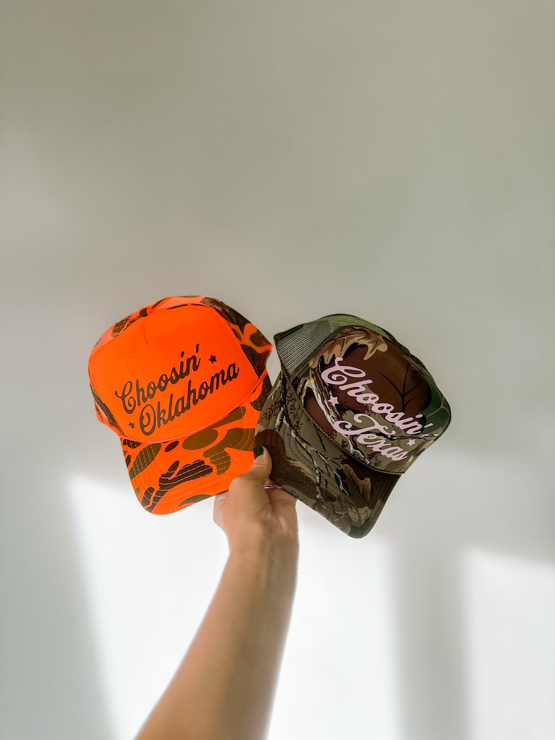 May include: Two baseball caps are held up against a white background. One is bright orange with the text "Choosin' Oklahoma" in brown script. The other is camouflage with the text "Choosin' Texas" in white script.