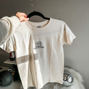 May include: A white t-shirt with a black graphic of two cowboy boots on the front.