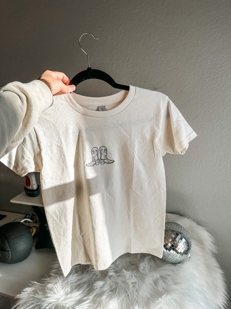 May include: A white t-shirt with a black graphic of two cowboy boots on the front.