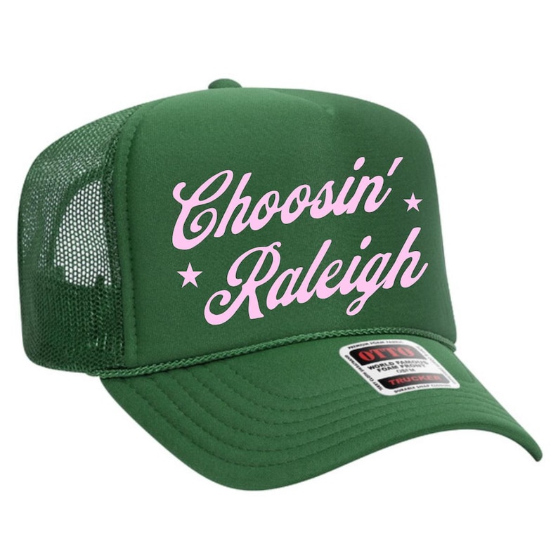May include: A green trucker hat with a mesh back and a curved brim. The hat features the text "Choosin' Raleigh" in pink script with star accents. The hat has a structured front panel and a snap closure.
