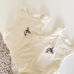 May include: Two cream-coloured vest tops with a brown embroidered design of a person riding a fish. The vests are made of a soft material and are laid on a white surface.