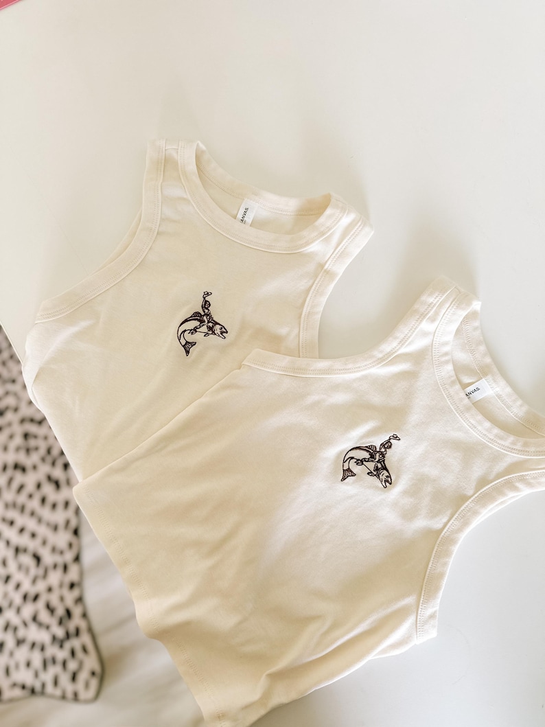 May include: Two cream-coloured vest tops with a brown embroidered design of a person riding a fish. The vests are made of a soft material and are laid on a white surface.