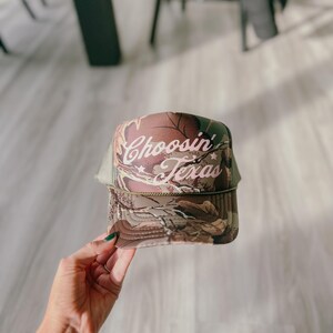 May include: A trucker hat with a camouflage pattern on the front panel and a mesh back. The hat features the text "Choosin' Texas" in a cursive font. The hat is a neutral color with a curved brim.