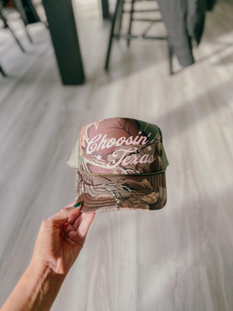 May include: A trucker hat with a camouflage pattern on the front panel and a mesh back. The hat features the text "Choosin' Texas" in a cursive font. The hat is a neutral color with a curved brim.