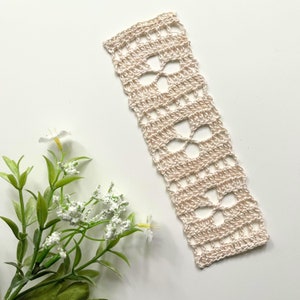 May include: A delicate cream-colored crocheted bookmark with a repeating floral pattern.