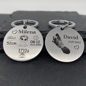 Può includere: Due portachiavi d'argento con dettagli incisi. Un portachiavi presenta un'impronta di piede di bambino, il nome "David" e la data "13.07.2022". L'altro portachiavi presenta un bambino, un cuore, un orologio, un metro a nastro, una bilancia, il nome "Milena", la data "03.11.2020", l'ora "08:12", il peso "3710g" e la lunghezza "52cm".