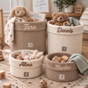 Op de afbeelding: Vier cilindervormige gebreide opbergmanden in beige en crème, elk voorzien van een op de rand geborduurde naam: Tara, Dennis, Alina en Marlon. Elke mand heeft een klein vierkant label met de letter J en bevat diverse knuffels.