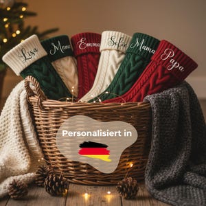 Könnte beinhalten: Ein Weidenkorb enthält sechs personalisierte Weihnachtsstrümpfe in Creme, Grün und Rot. Jeder Strumpf ist mit einem Namen in weißer Schrift bestickt. Die Szene zeigt eine Decke, Tannenzapfen und den Text 'Personalisiert in' mit einer deutschen Flagge.