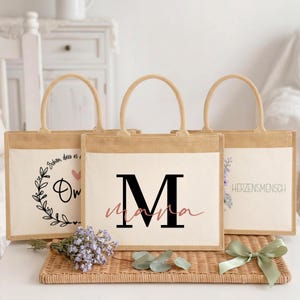 Könnte beinhalten: Drei rechteckige Jute-Tragetaschen mit cremefarbenen Canvas-Einsätzen. Die mittlere Tasche zeigt ein großes schwarzes M mit dem Wort mama in rosa Schreibschrift. Die linke Tasche hat einen Blumenkranz und ein Herz, die rechte Tasche zeigt den Text HERZENSMENSCH.