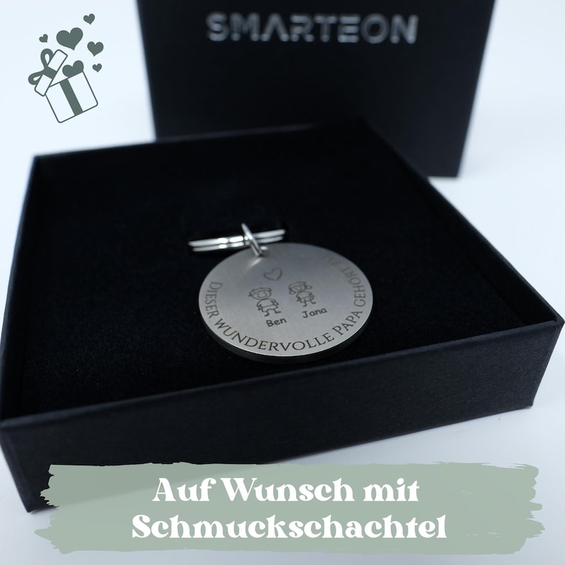 May include: A silver keychain with a round charm engraved with the text "Dieser wundervolle Papa gehoert..." and two stick figures of children with the names "Ben" and "Jana" below. The keychain is in a black gift box with the text "Auf Wunsch mit Schmuckschachtel" on a green background.