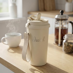 Op de afbeelding: Een crèmekleurige reismok met een klapdeksel en een bijpassende stoffen band. De naam "Isabel" staat in sierlijke letters op de zijkant. Een witte theekop en een cafetière staan op de achtergrond.