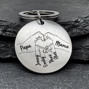 May include: Silver keychain with a heart formed by two hands, with the words "Papa" and "Mama" on either side. Inside the heart are four smaller hands with the names "Jenny", "Emily", "Theo", and "Alex" written below.