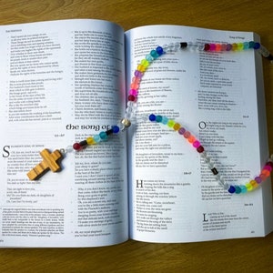 May include: An open book with a wooden cross and a colourful beaded necklace on top. The book is open to the Song of Songs and the book of Psalms.