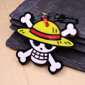 Peut inclure: Un porte-clés jaune, rouge et noir représentant un crâne et des os croisés avec un chapeau de paille. Le porte-clés est doté d'un fermoir noir et d'une ficelle rouge pour l'attache. Le design est un symbole populaire d'anime et de manga.