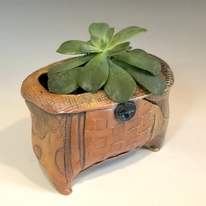 Puede incluir: Un pequeño macetero de cerámica con un acabado texturizado marrón y beige. El macetero tiene una forma única, asimétrica con cuatro patas y un pequeño pomo redondo en el lateral. Una planta suculenta verde está creciendo en el macetero.