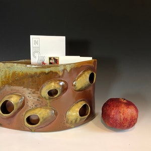 Pode incluir: Um porta-cartas de cerâmica artesanal em tons de castanho e bege, com seis aberturas circulares. Contém envelopes brancos com selos. Uma romã vermelha está colocada ao lado.