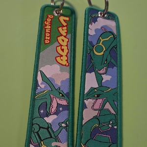 May include: Two rectangular embroidered keychains featuring the green serpentine Pokémon Rayquaza against a blue sky with white clouds. One keychain includes the text Rayquaza and Japanese characters in red and yellow. Both have silver metal rings.