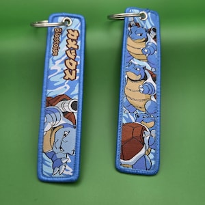 May include: Two rectangular embroidered keychains featuring the blue Pokémon Blastoise. One keychain displays the Japanese text カメックス and English text Blastoise, while the other shows multiple Blastoise figures. Both have blue borders and metal rings.