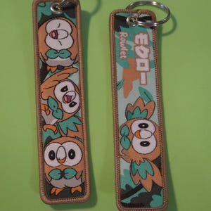 May include: Two rectangular fabric keychains with embroidered edges. The left keychain features three stacked Rowlet characters. The right keychain displays one Rowlet character with the text "モクロー" and "Rowlet" printed vertically in white.