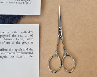 May include: A pair of small, gold-colored scissors with ornate designs on the handles. The scissors are open and the blades are sharp. The scissors are on a brown surface.