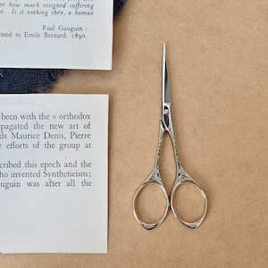 May include: A pair of small, gold-coloured scissors with ornate designs on the handles. The scissors are open and the blades are sharp. The scissors are on a brown surface.