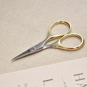 May include: Small gold and silver scissors with a pointed tip. The scissors have two loops, one gold and one silver.