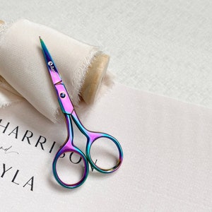 May include: Multi-coloured iridescent scissors with a pointed tip and rounded finger holes. The scissors are on white fabric with the text "HARRISON" and "YLA". The scissors have a small logo on the blade.