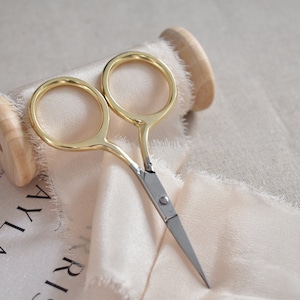 May include: Gold-toned small scissors with silver blades, resting on a spool of pale pink ribbon and a white paper with black text that reads "SATURDAY 7TH".