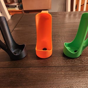 May include: Three 3D-printed holders in black, orange, and green. Each has a curved handle and a small, open base. The orange holder is centered, flanked by the black and green ones. They are displayed on a dark wood surface.
