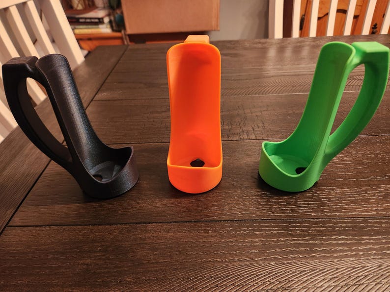 May include: Three 3D-printed holders in black, orange, and green. Each has a curved handle and a small, open base. The orange holder is centered, flanked by the black and green ones. They are displayed on a dark wood surface.