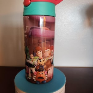 May include: A cylindrical water bottle featuring a colorful graphic print of various animated characters including a cowboy, a space ranger, a potato-shaped figure, and a horse. The bottle has a teal neck and a red lid with a carry handle.