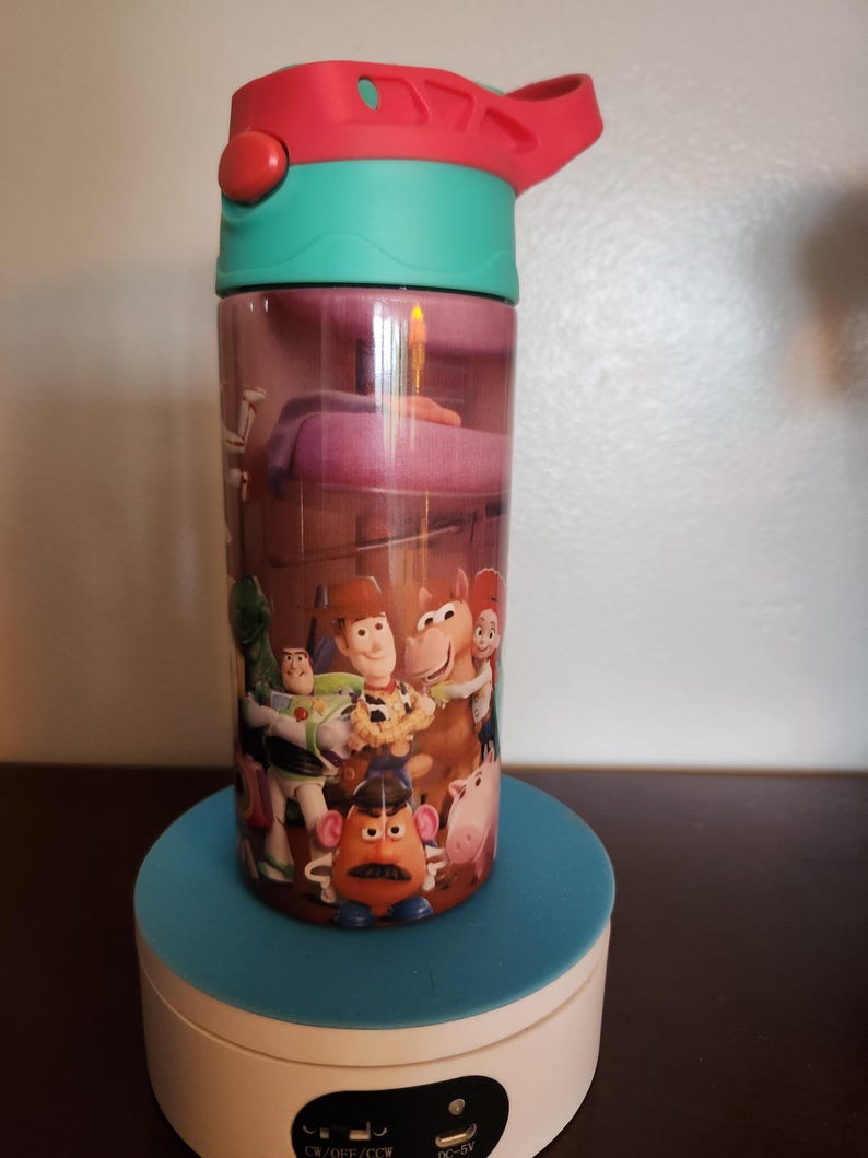 May include: A cylindrical water bottle featuring a colorful graphic print of various animated characters including a cowboy, a space ranger, a potato-shaped figure, and a horse. The bottle has a teal neck and a red lid with a carry handle.