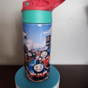 May include: A stainless steel water bottle featuring a colorful design of Thomas the Tank Engine and other train characters. The bottle has a teal lid and a red handle. The bottle is cylindrical and sits on a blue rotating stand.