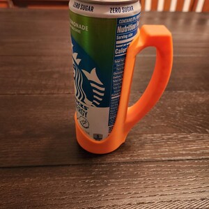 May include: An orange plastic handle attached to a Starbucks energy drink can. The can is green and white with the Starbucks logo and the words "ZERO SUGAR" and "MONADE". The handle is designed to make the can easier to hold.