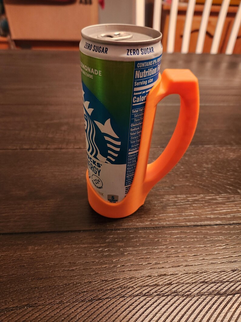 May include: An orange plastic handle attached to a Starbucks energy drink can. The can is green and white with the Starbucks logo and the words "ZERO SUGAR" and "MONADE". The handle is designed to make the can easier to hold.