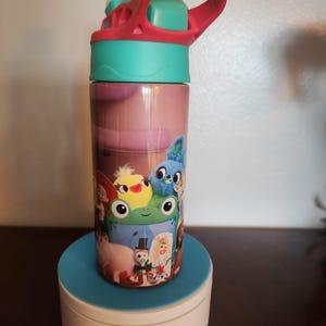 May include: A cylindrical water bottle featuring a colorful graphic print of various animated characters, including a green frog, a yellow chick, and a blue creature. The bottle has a bright teal lid with a pink carrying handle and a clear straw.