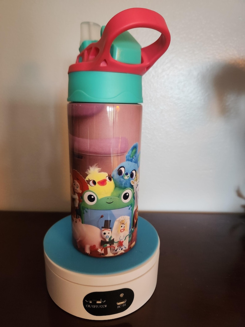 May include: A cylindrical water bottle featuring a colorful graphic print of various animated characters, including a green frog, a yellow chick, and a blue creature. The bottle has a bright teal lid with a pink carrying handle and a clear straw.