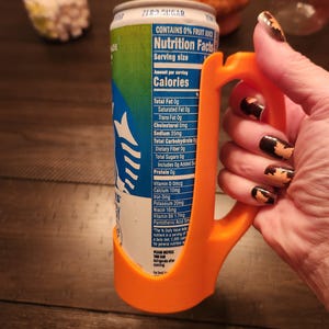 May include: An orange 3D-printed can holder with a handle, wrapped around a can of a beverage. The can has a blue and green label with nutrition facts. The text "ZERO SUGAR" is visible on the can.