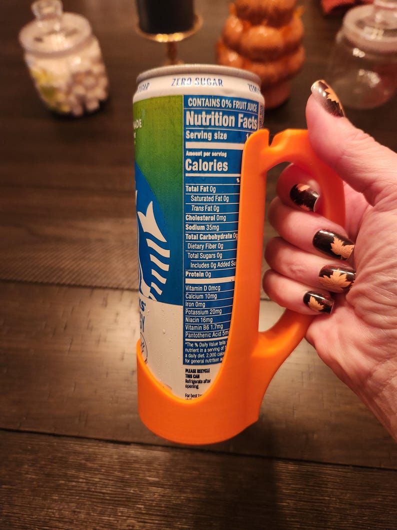 May include: An orange 3D-printed can holder with a handle, wrapped around a can of a beverage. The can has a blue and green label with nutrition facts. The text "ZERO SUGAR" is visible on the can.