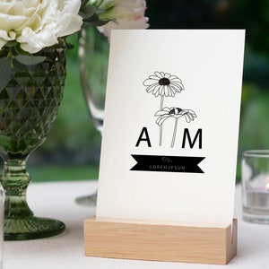 May include: A white card with black line drawings of daisies and the letters "A M" with a black ribbon that says "The Lorem Ipsum" below. The card is on a wooden stand.