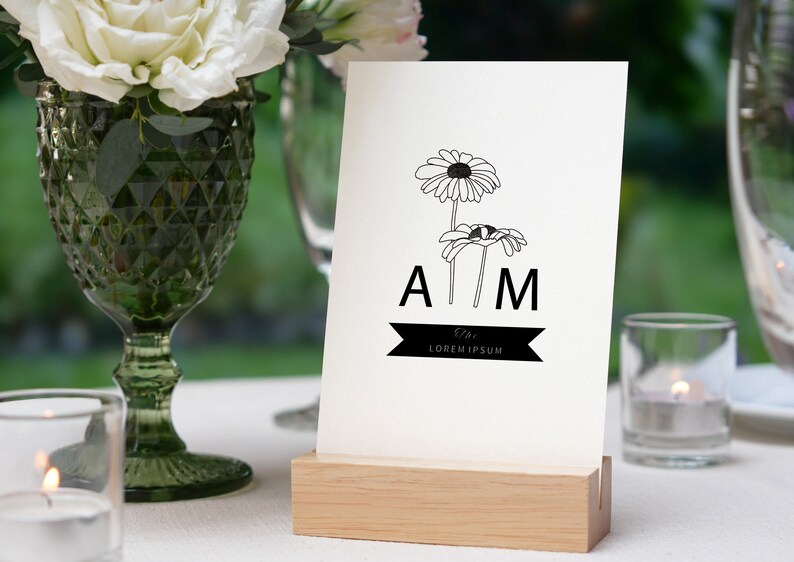 May include: A white card with black line drawings of daisies and the letters "A M" with a black ribbon that says "The Lorem Ipsum" below. The card is on a wooden stand.