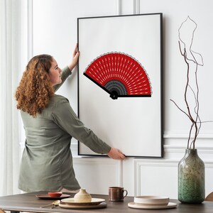 Puede incluir: Una mujer cuelga una impresi&oacute;n enmarcada de un abanico rojo y negro en una pared blanca. El abanico est&aacute; abierto y tiene un mango negro.