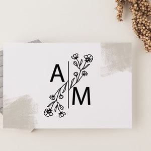 May include: A white card with a black floral design and the letters "A" and "M" on either side of the floral design. The card has a gray painted background.