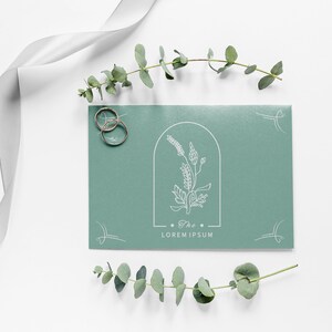 May include: A green invitation card with a white floral design and the text "The Lorem Ipsum" in a minimalist font. Two gold rings are placed on the left side of the card.