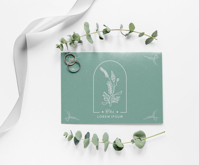 May include: A green invitation card with a white floral design and the text "The Lorem Ipsum" in a minimalist font. Two gold rings are placed on the left side of the card.