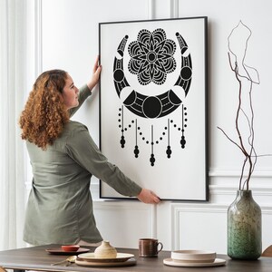May include: A person hangs a black and white framed print of a moon and flower design on a white wall. The print is in a modern style and features a crescent moon with a flower in the center. The print is surrounded by a thin black frame.
