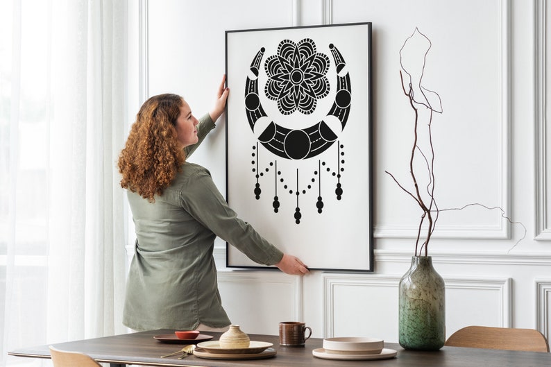 May include: A person hangs a black and white framed print of a moon and flower design on a white wall. The print is in a modern style and features a crescent moon with a flower in the center. The print is surrounded by a thin black frame.