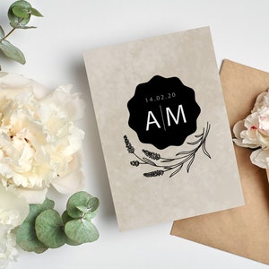 May include: A wedding invitation with a simple design featuring a black circle with the date "14.02.20" and the initials "A M" in white. The circle is surrounded by a floral design.